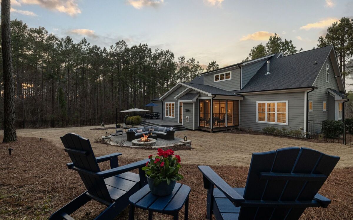 Modern residential backyard at dusk with a fire pit, Adirondack chairs, paved terrace and house beyond